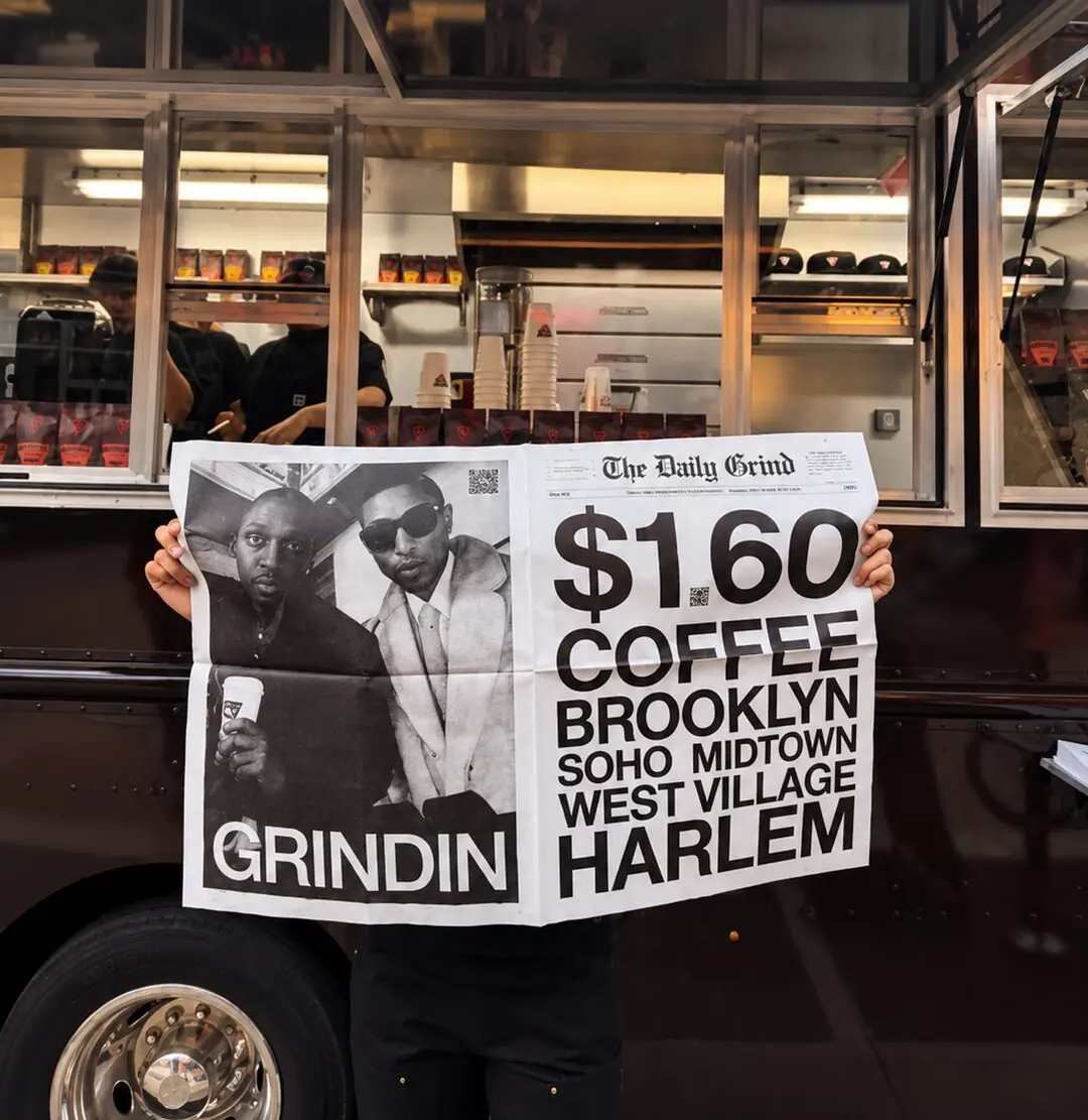 A Grindin crew member holding up a two-page newspaper spread promoting $1.60 coffee in Brooklyn, SoHo, Midtown, West Village and Harlem, in front of the wrapped Grindin truck