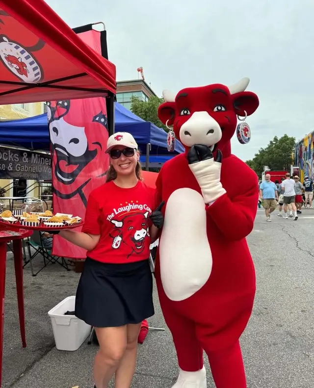 A Laughing Cow sampling team member in branded red gear posing with the brand's mascot beside trays of samples
